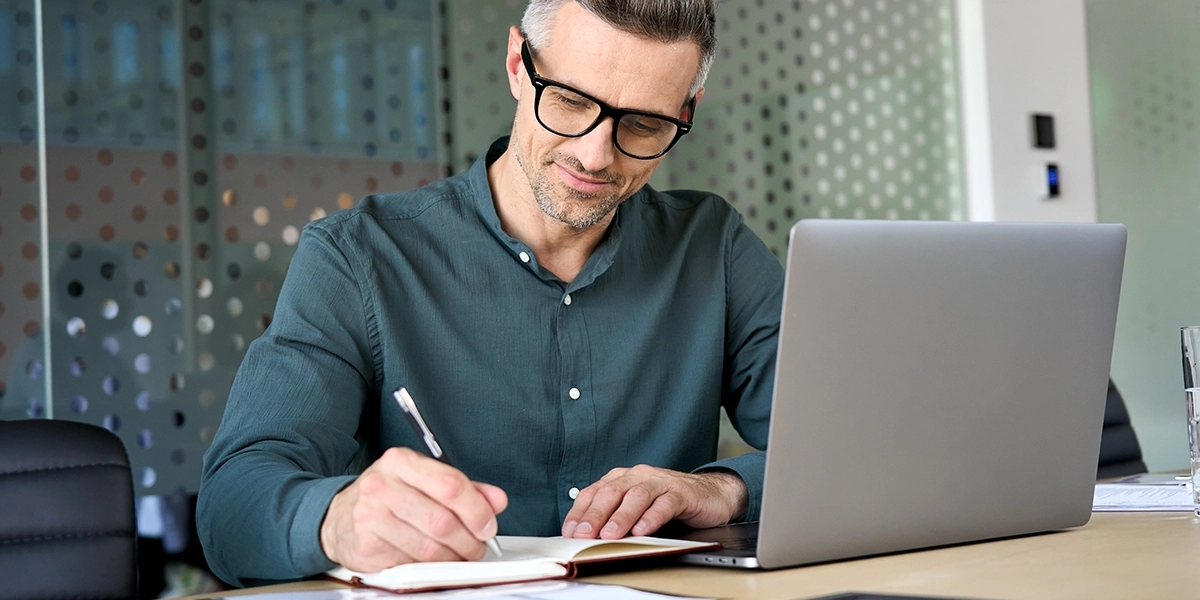 A man writes in his notebook with his laptop nearby.