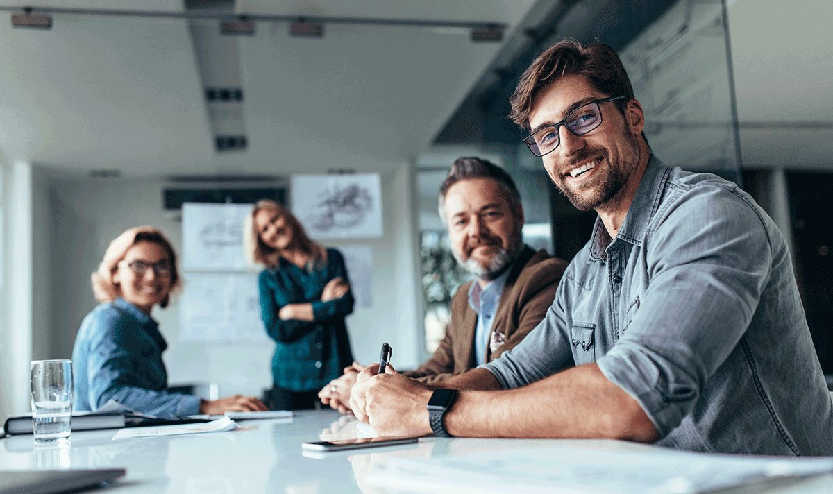 Four smiling people, two women and two men, are sitting or standing around a conference table in a business office.