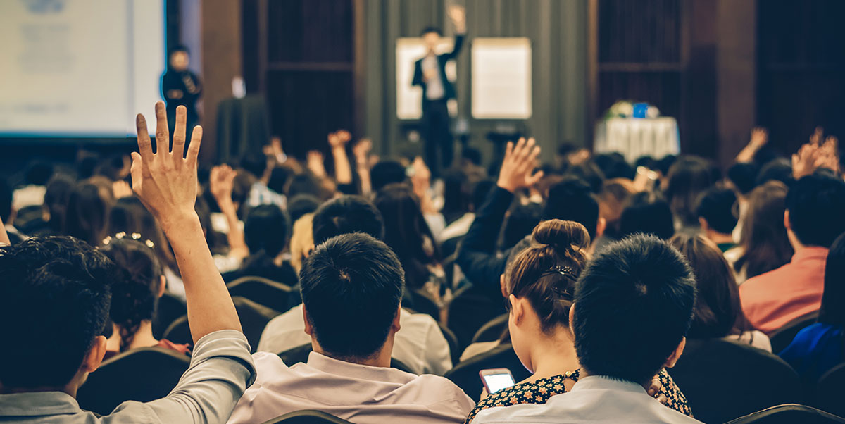 At a conference, a speaker stands on stage, and people in the audience are raising their hands to be called on.
