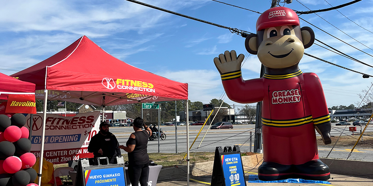 A Grease Monkey mascot is beside a vendor booth at a Grease Monkey grand opening.